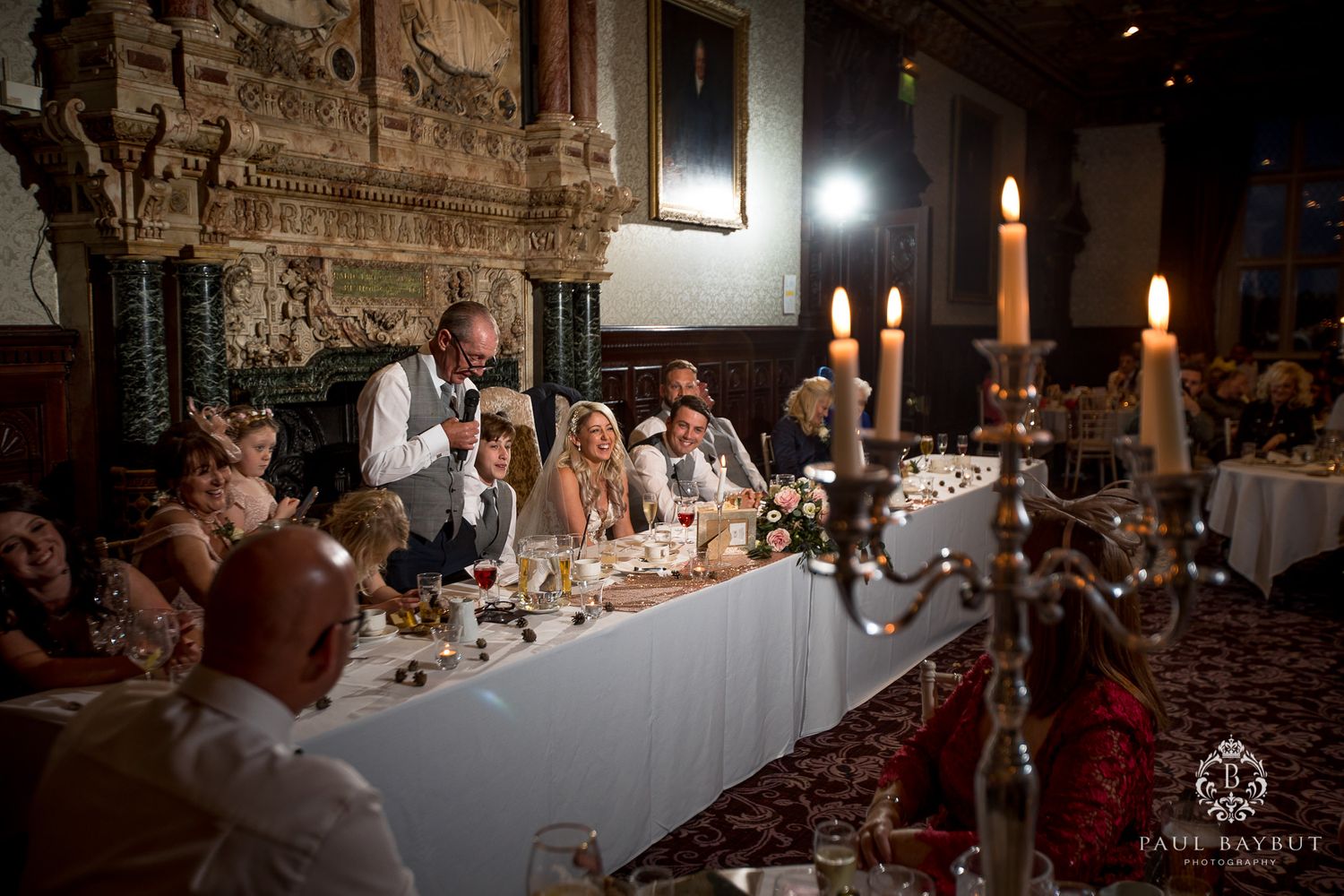 Candlelit wedding reception with guests gathered at long tables in an ornate historic hall.