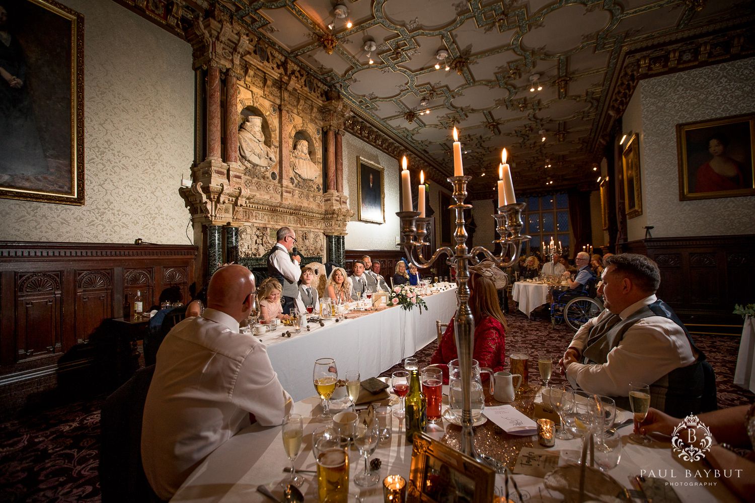 Long banquet table set for wedding reception in historic hall with ornate decor and candlelight.