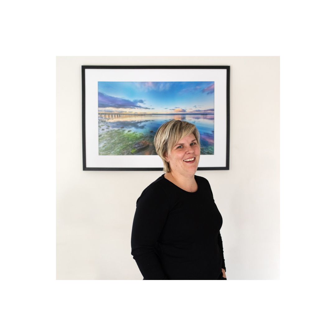 Coastal seascape photograph hanging on white wall with person standing in front.