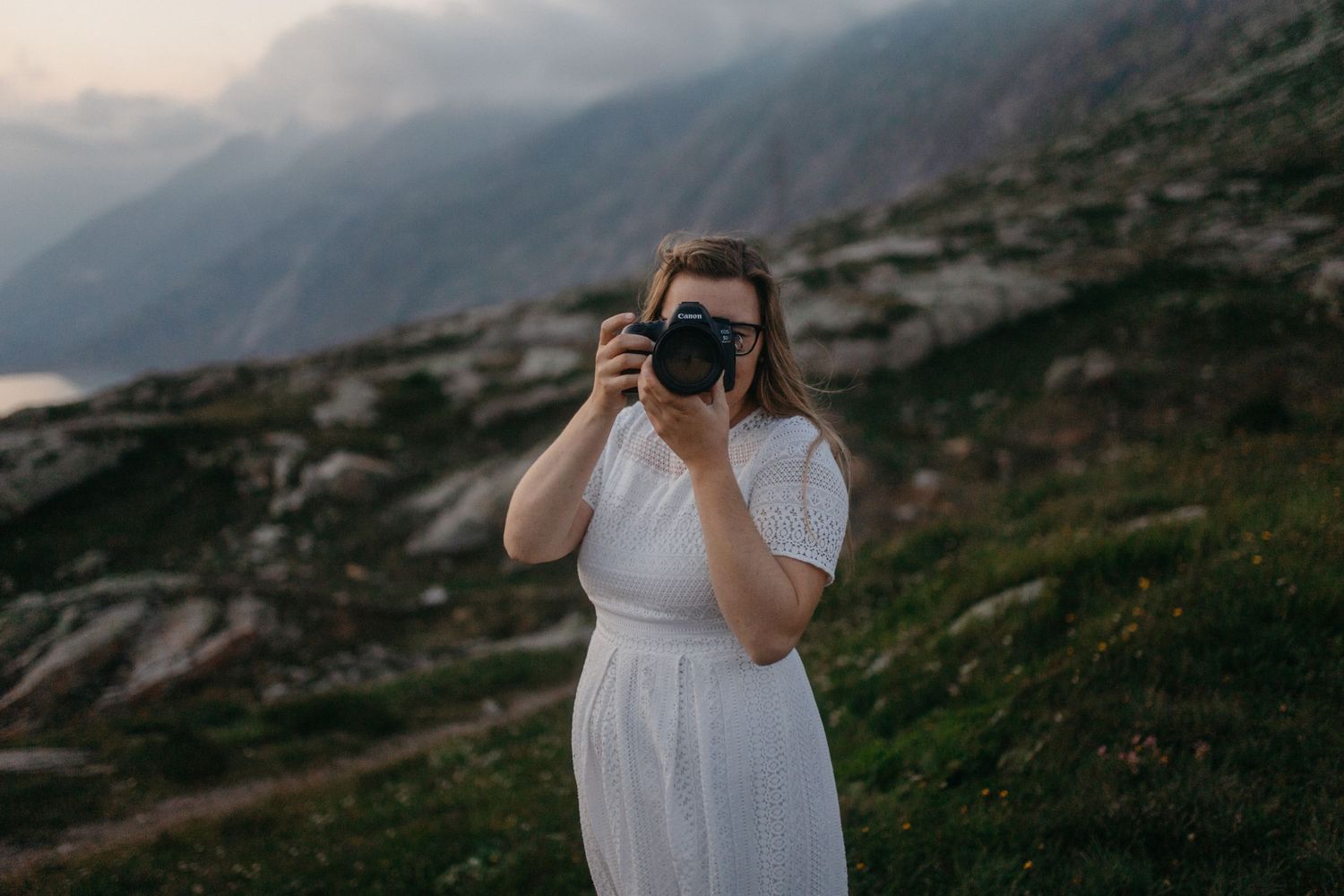 A person in white dress takes photographs in a mountainous landscape at dusk.
