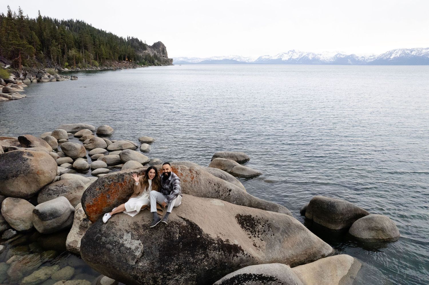 A large dog rests on rocky beach boulders overlooking Lake Tahoe with snow-capped mountains in the distance.