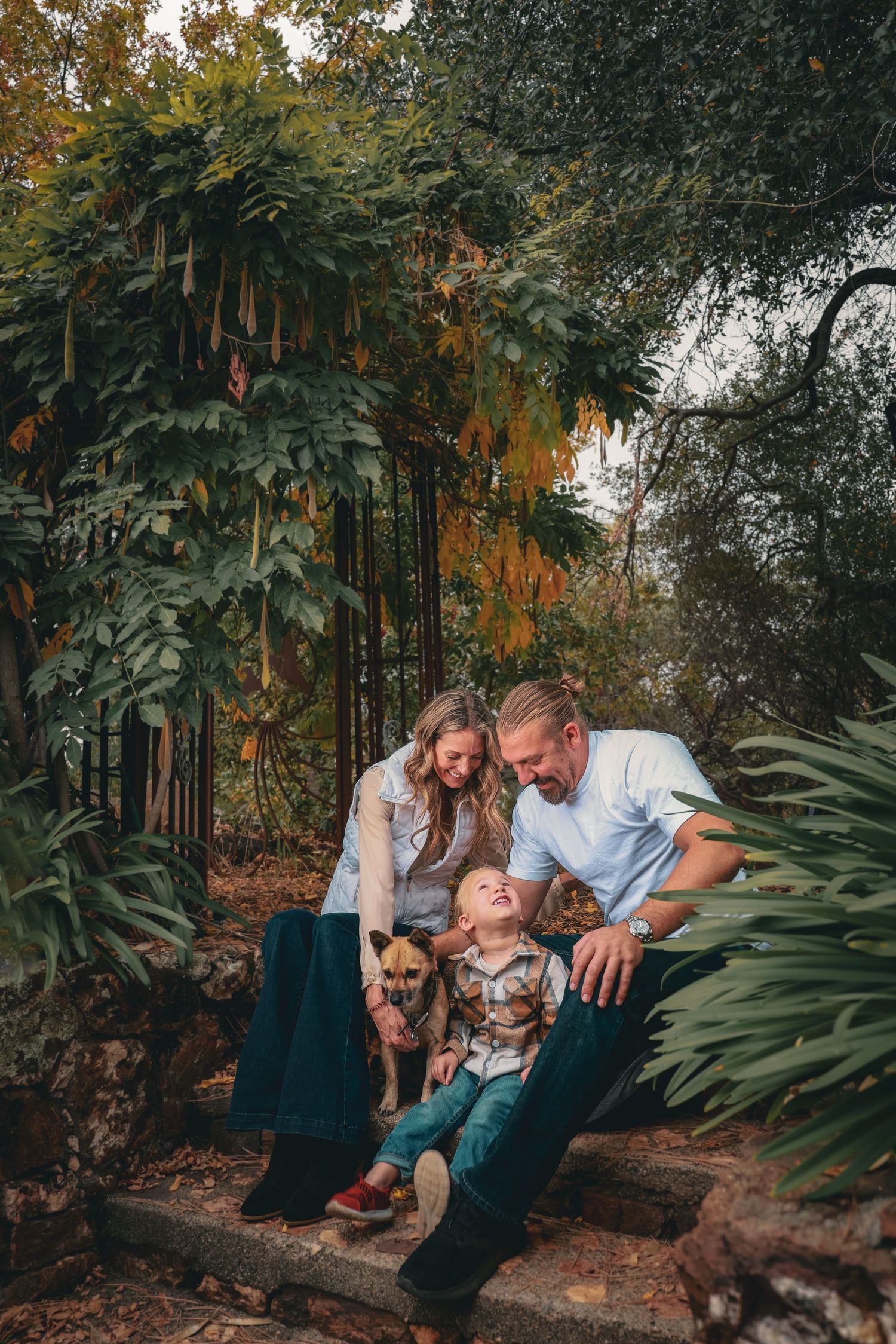 Happy family with child and small dog sitting on stone steps surrounded by lush green tropical garden foliage.