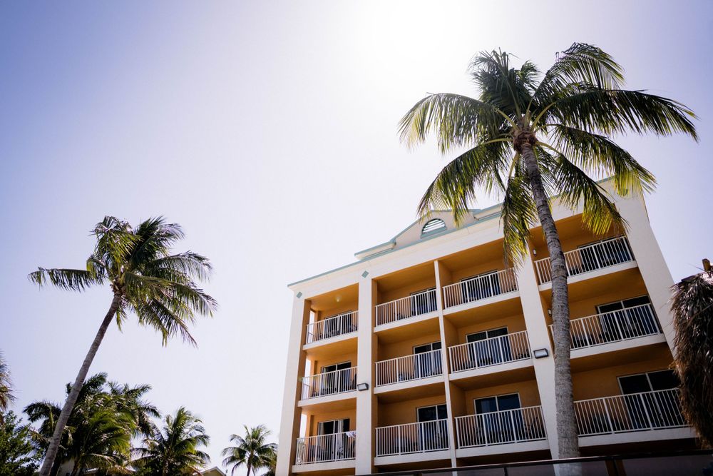 Modern beachfront hotel with yellow facade and multiple balconies surrounded by swaying palm trees.