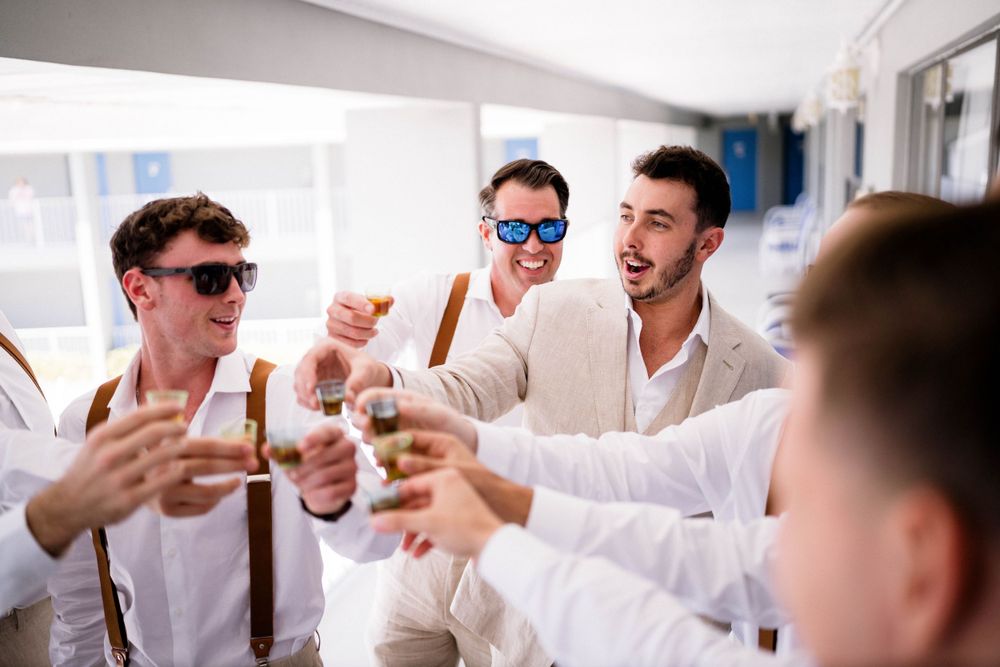Groomsmen in white shirts and sunglasses toast with drinks during wedding preparations.