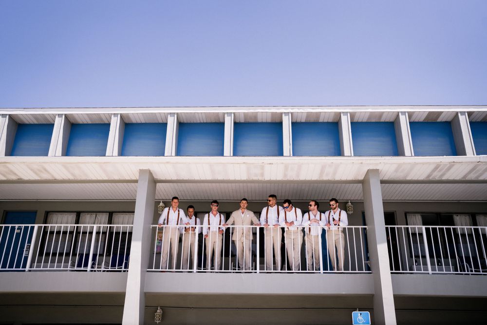 Wedding party in light colored attire poses together on a white balcony overlooking the ocean.