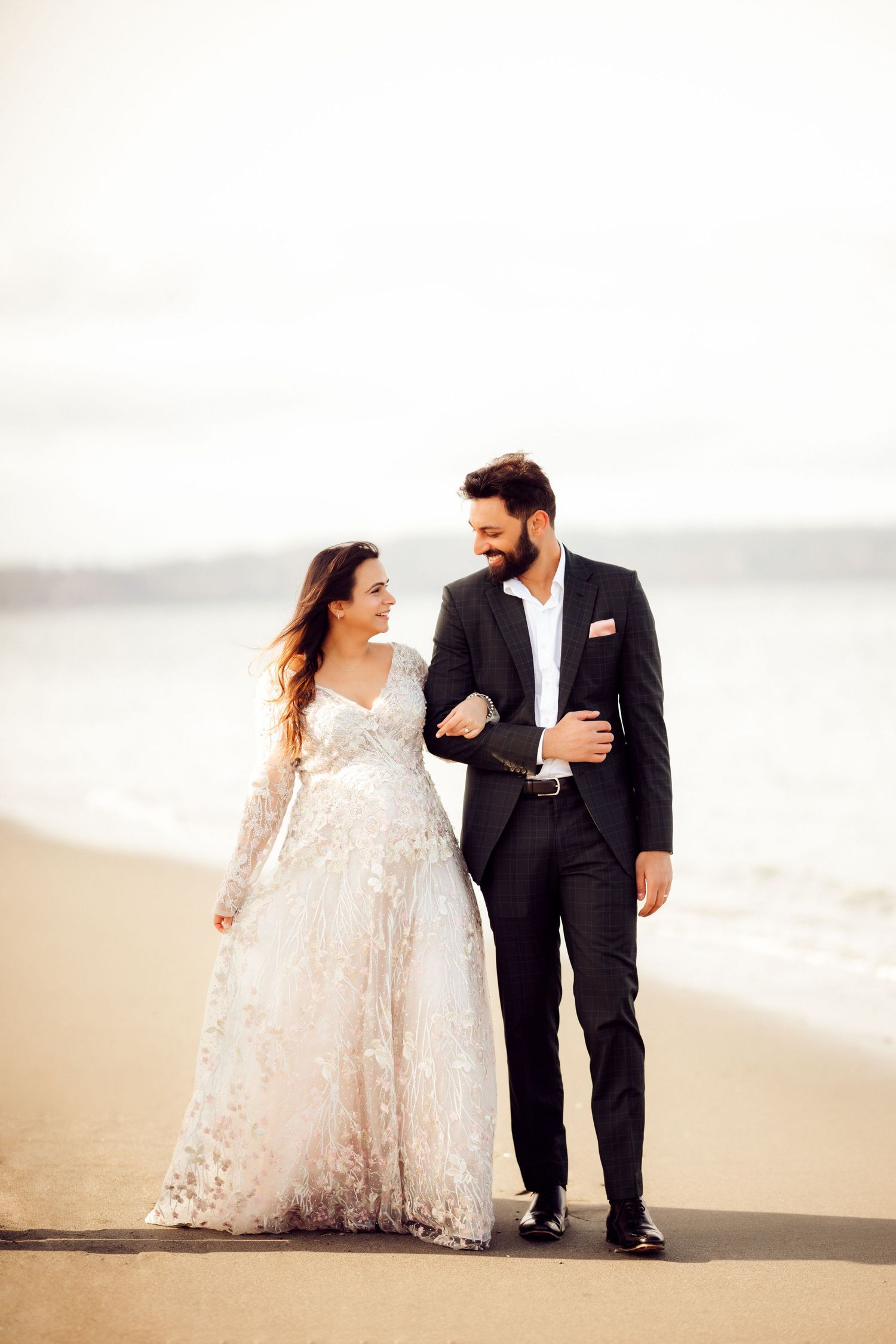 A couple walks together on a beach, bride in lace dress and groom in dark suit.