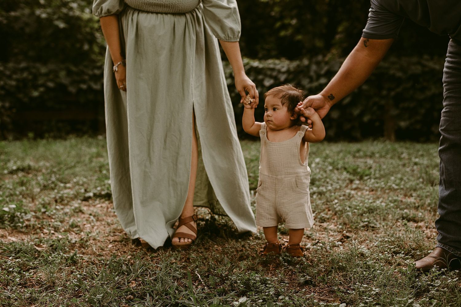 Close-up of parents helping small child take first steps on grassy lawn.