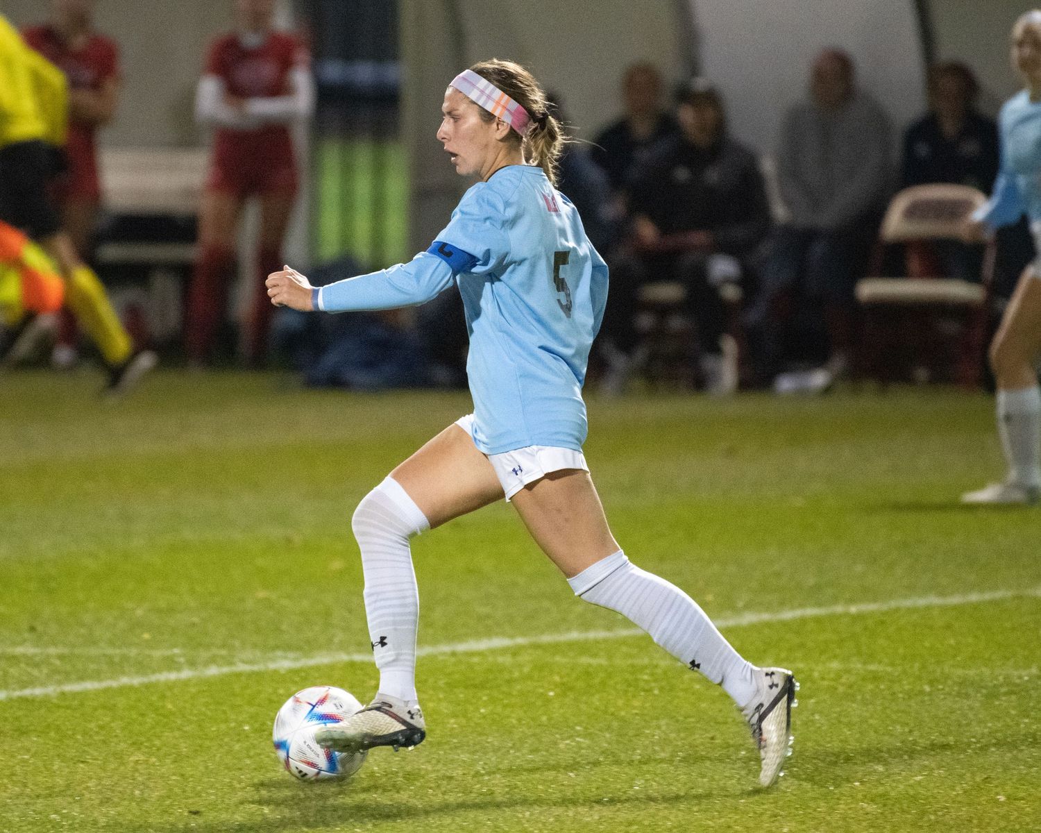Soccer player in light blue jersey dribbling ball during evening match on grass field.