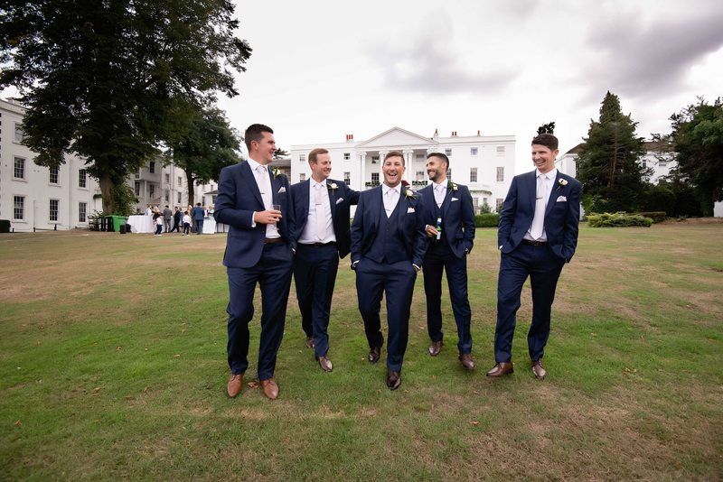Groomsmen in navy suits share a laugh while walking on a stately manor lawn.