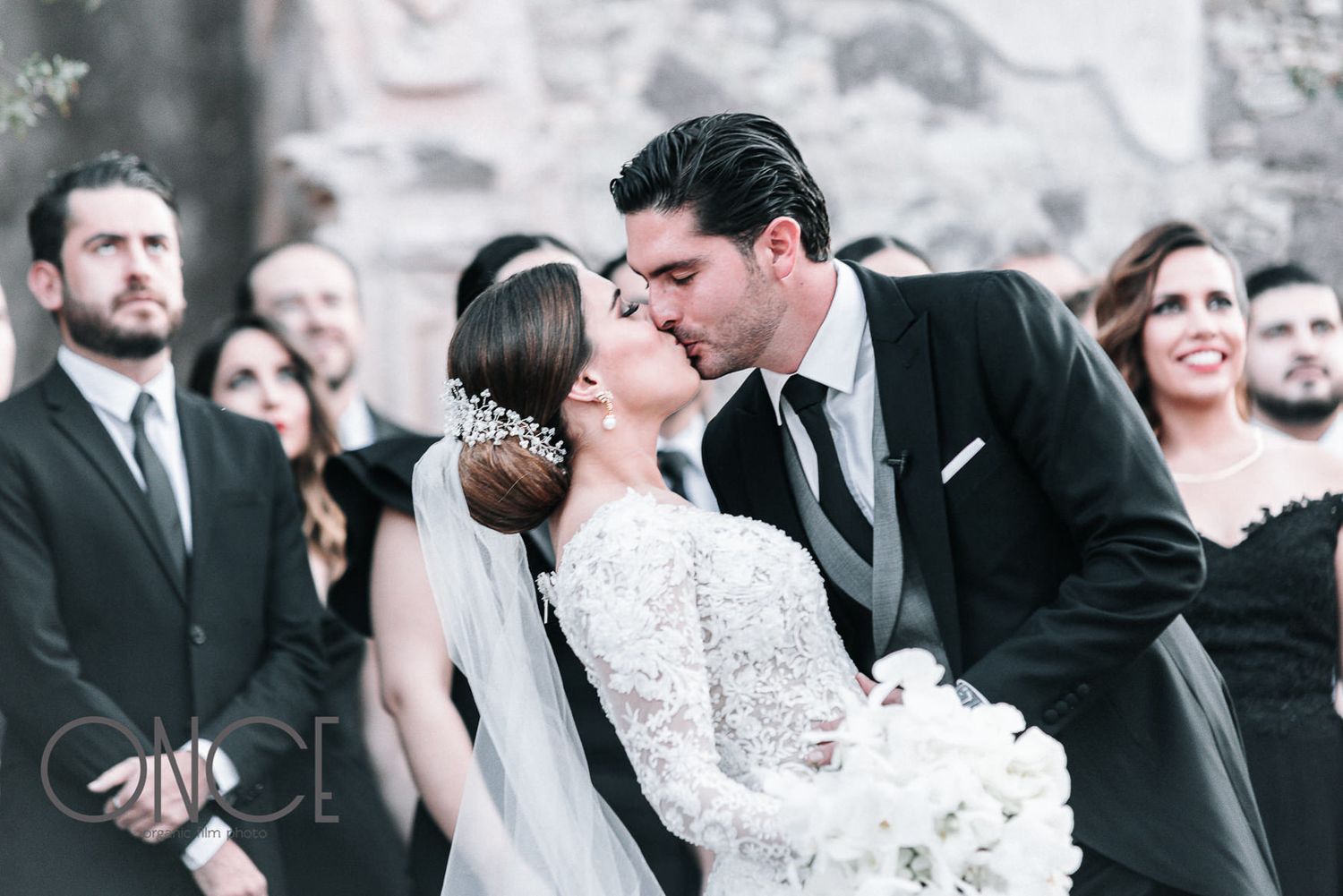 An intimate wedding ceremony kiss captured in black and white with guests looking on.
