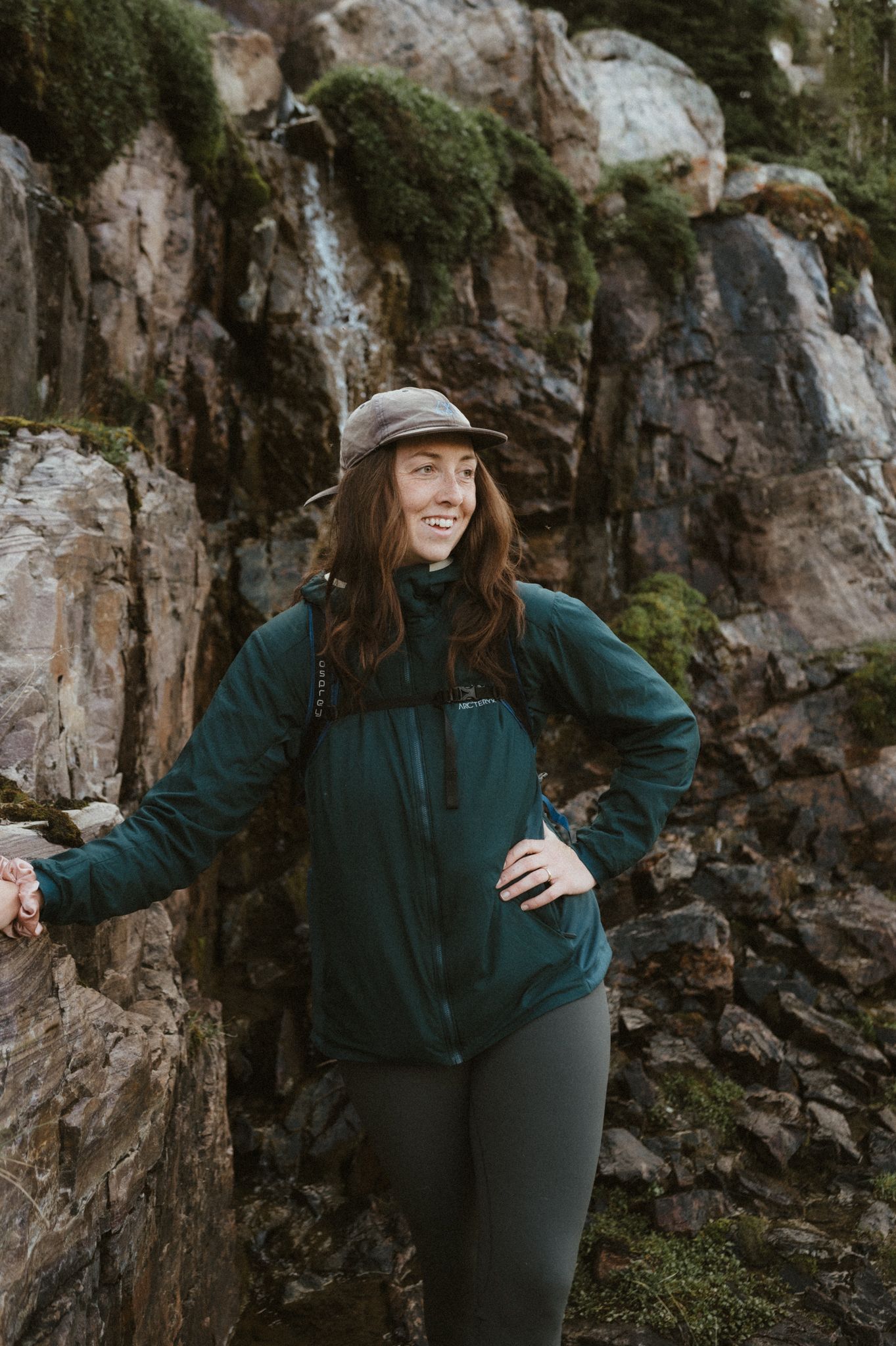 Outdoor enthusiast poses against mossy rock face wearing teal hiking jacket and baseball cap.