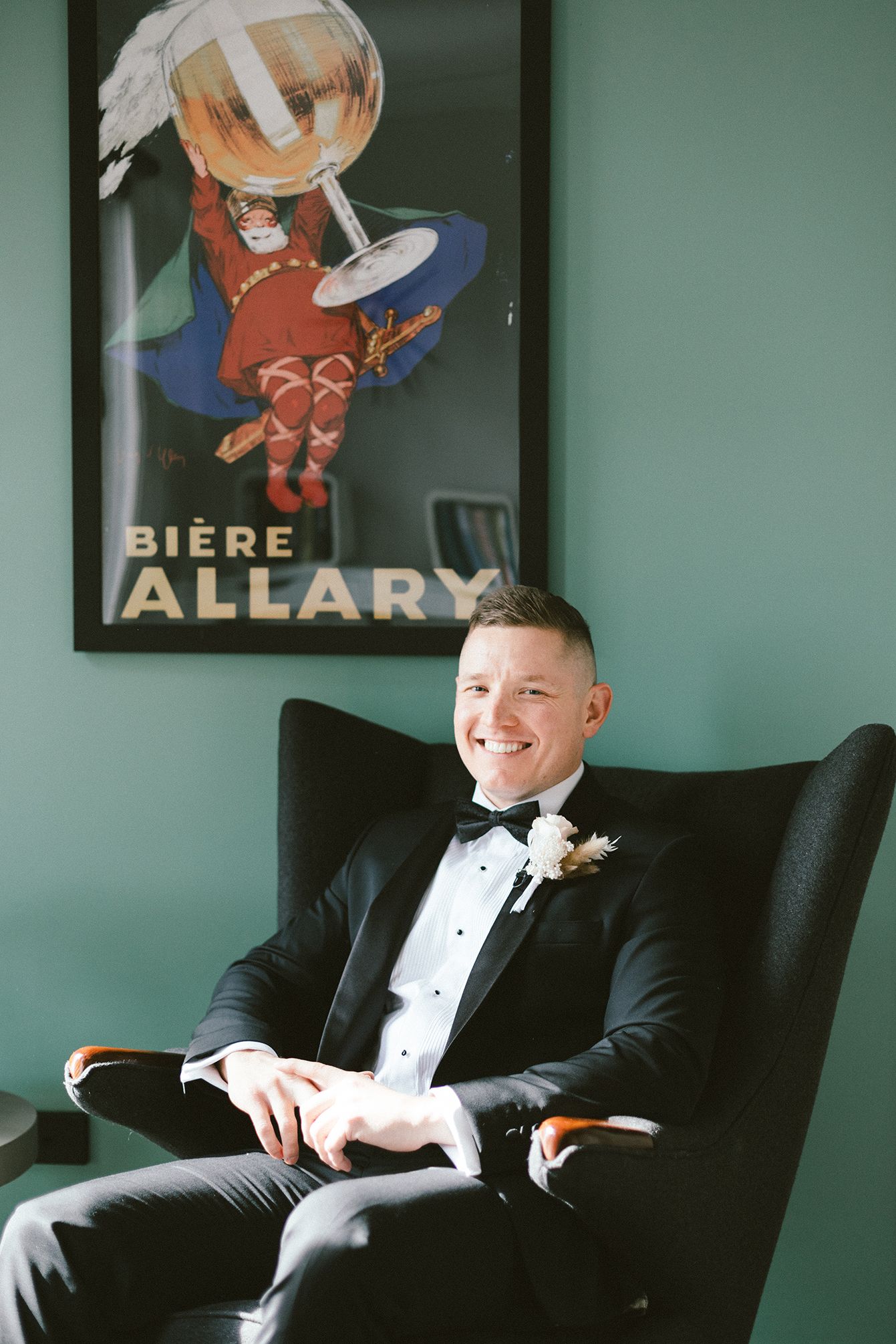 A person in formal attire sits in a dark chair against a green wall with vintage Biere Allary poster.