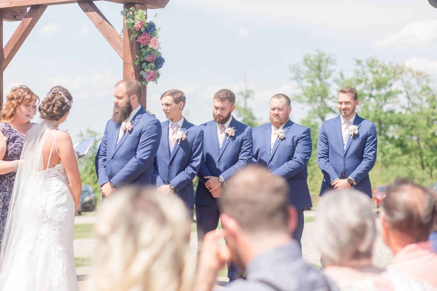 Wedding party members in navy suits and formal attire stand together during an outdoor ceremony.