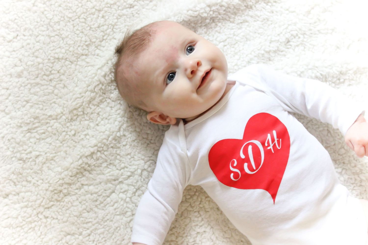 Baby wearing white onesie with red heart design lies on soft blanket.
