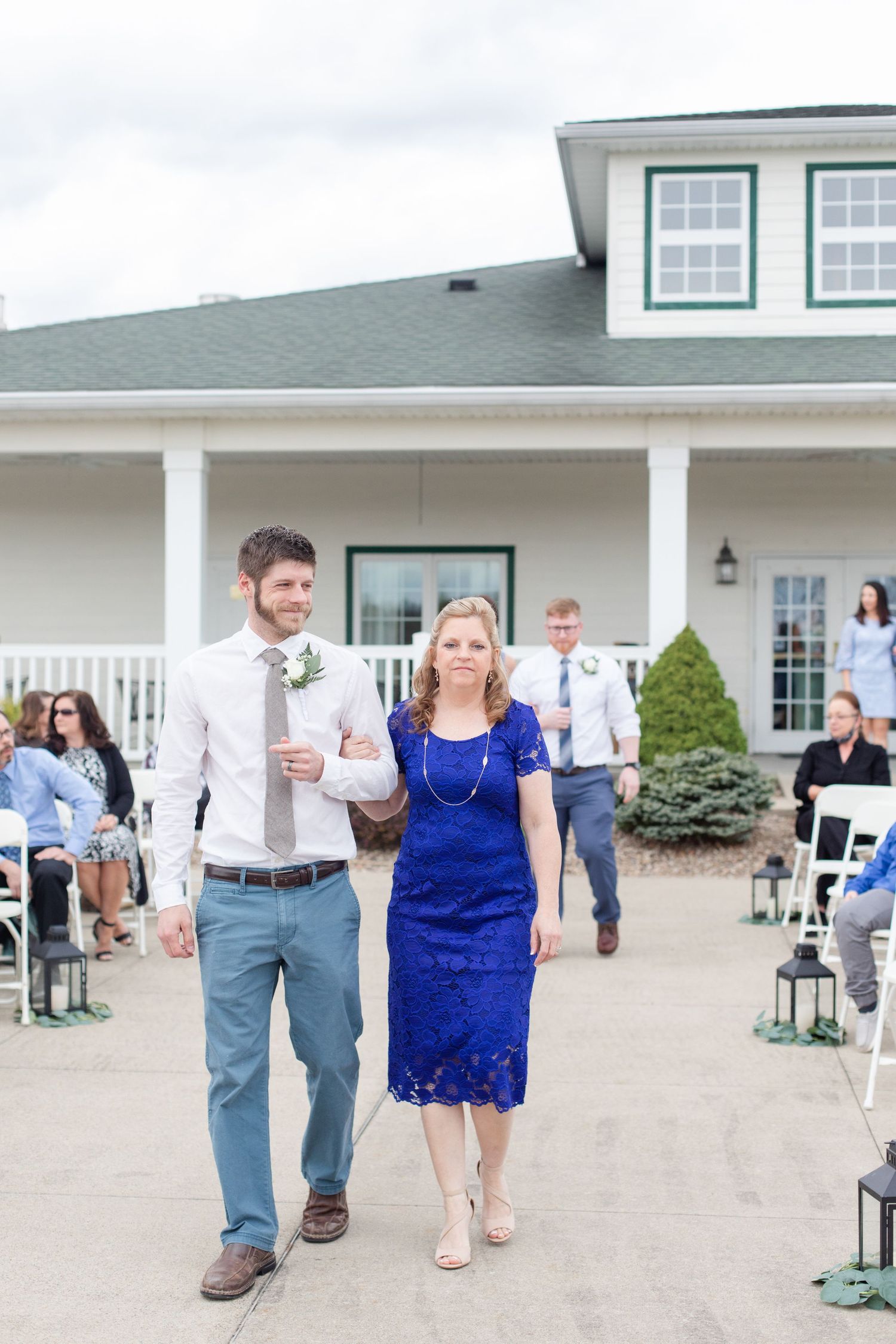 Wedding processional outside a white clubhouse with guests seated on folding chairs.