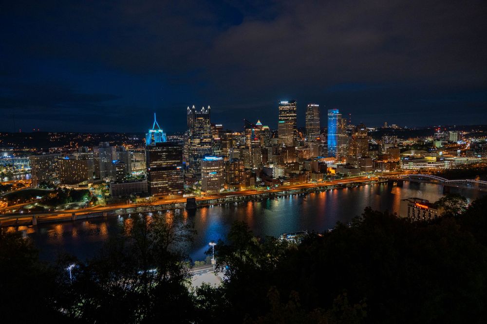 Pittsburgh city skyline at night with illuminated buildings reflecting in river.