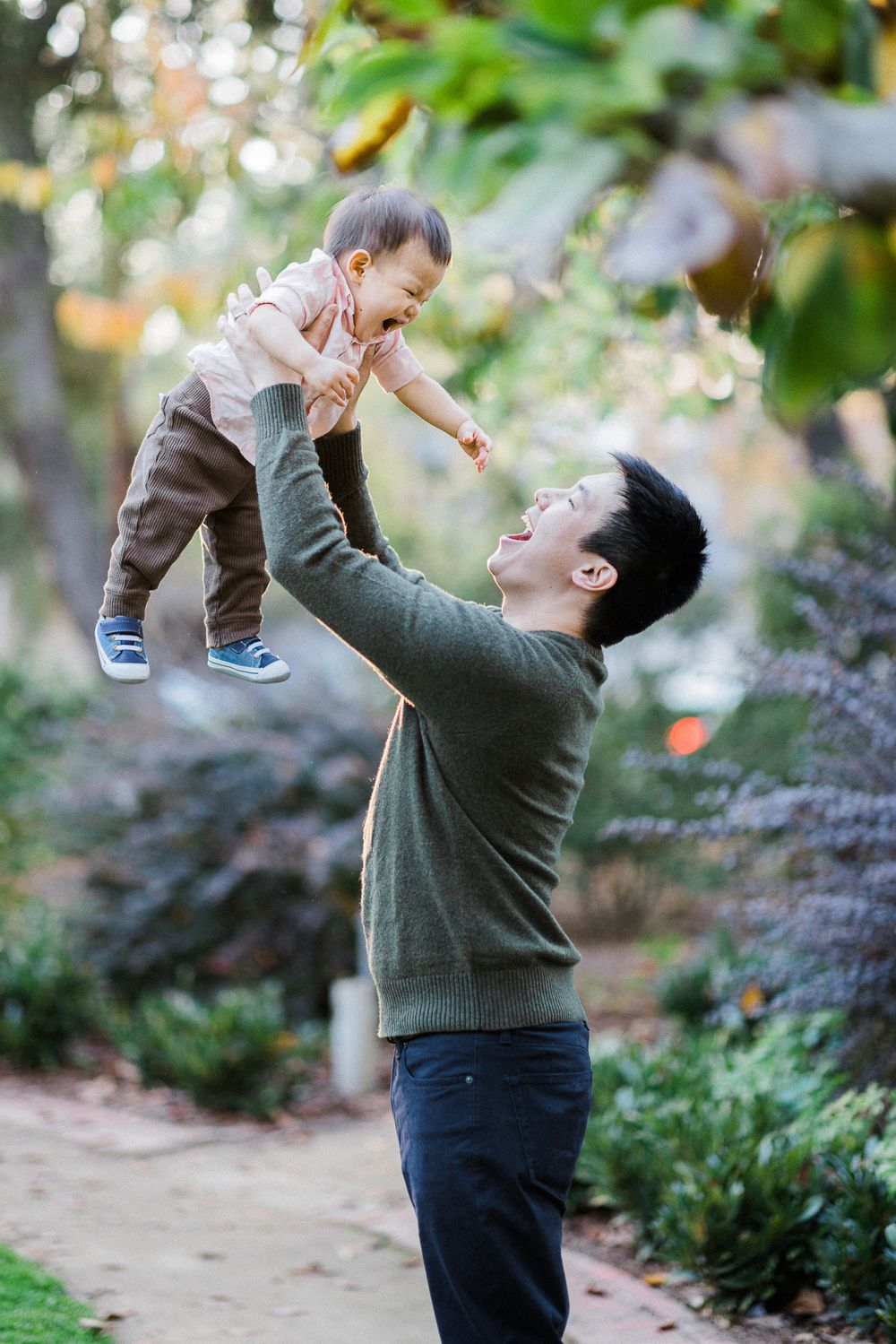 Parent lifts smiling baby high in the air while standing on garden path surrounded by greenery.
