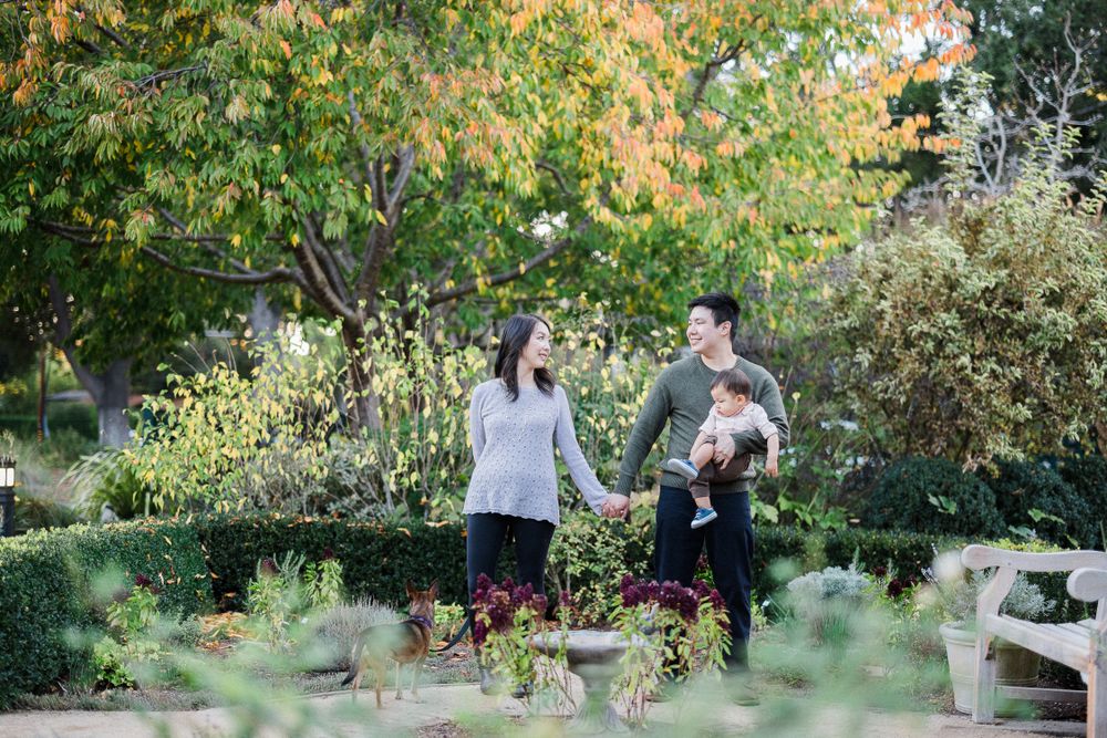 A family stands together in a lush garden setting with fall foliage in the background.