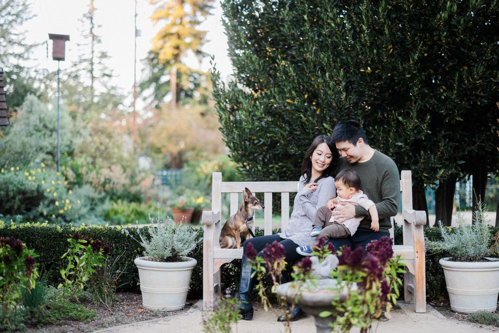 A couple sits with their baby on a garden bench surrounded by potted plants and greenery.