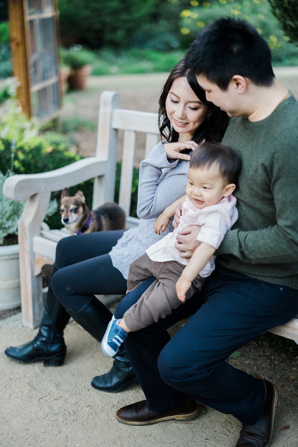 A family relaxes on a wooden bench in a garden setting with their small dog.