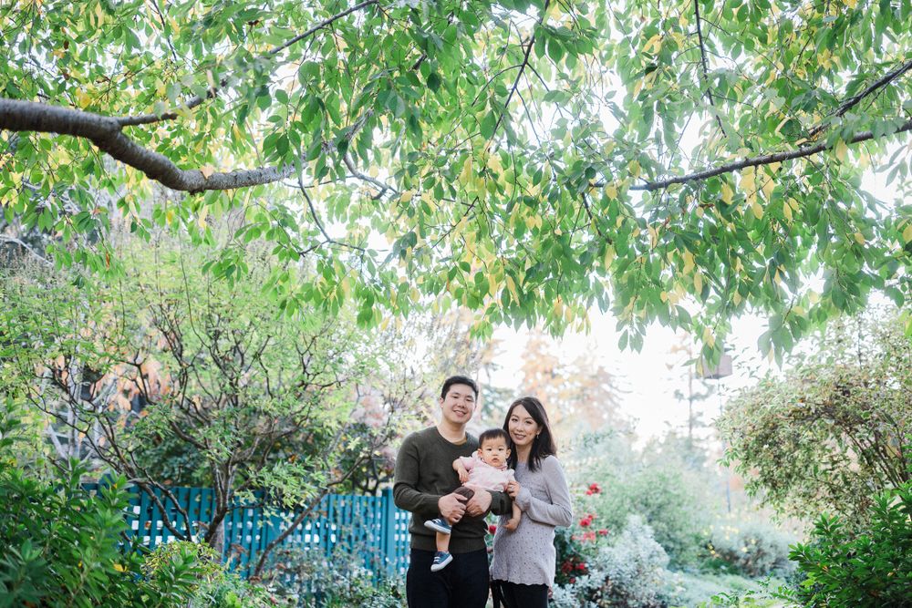 A family poses for photos in a lush garden with trees and blue fence in background.