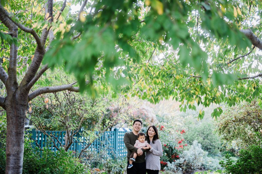 A family portrait captured under green trees with a blue fence in the background.