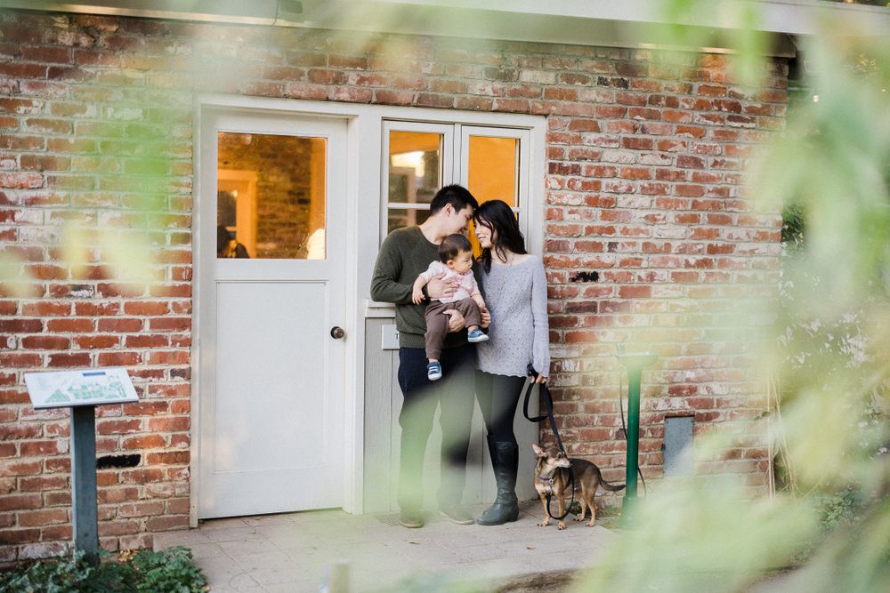 Family portrait taken at white doorway of brick building with greenery in foreground.