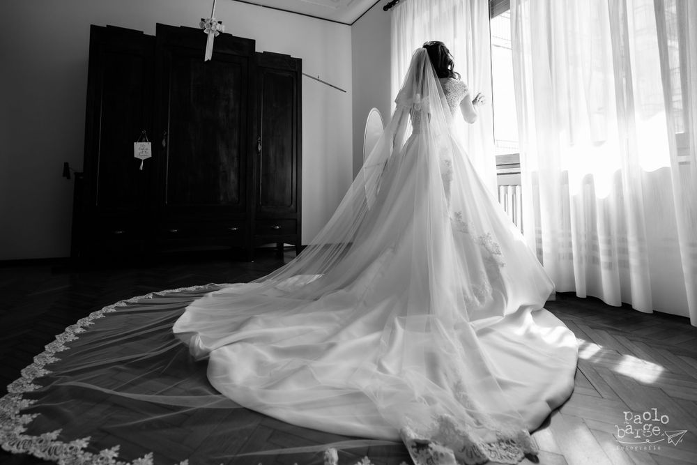 A black and white photo of a bride in a flowing wedding dress with a long train standing near a window.