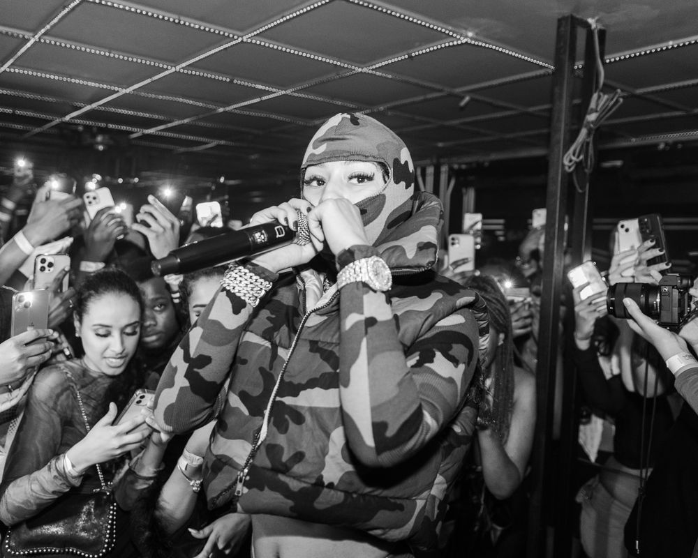 Black and white photo of masked performer in camo outfit rapping into microphone at crowded music venue.