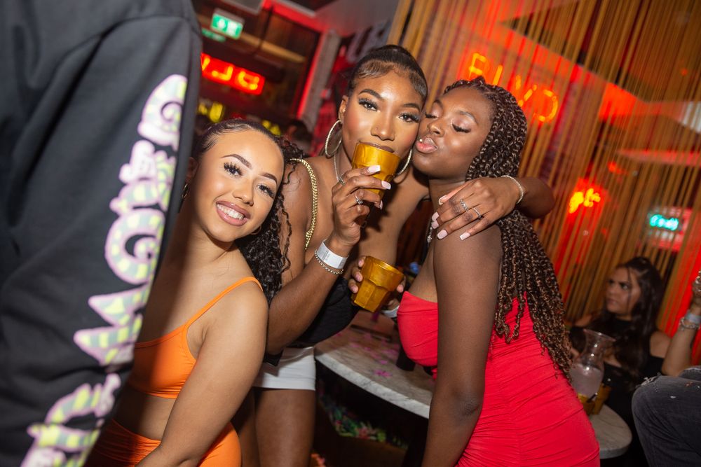 Three women enjoying drinks and posing together at a vibrant red-lit nightclub.