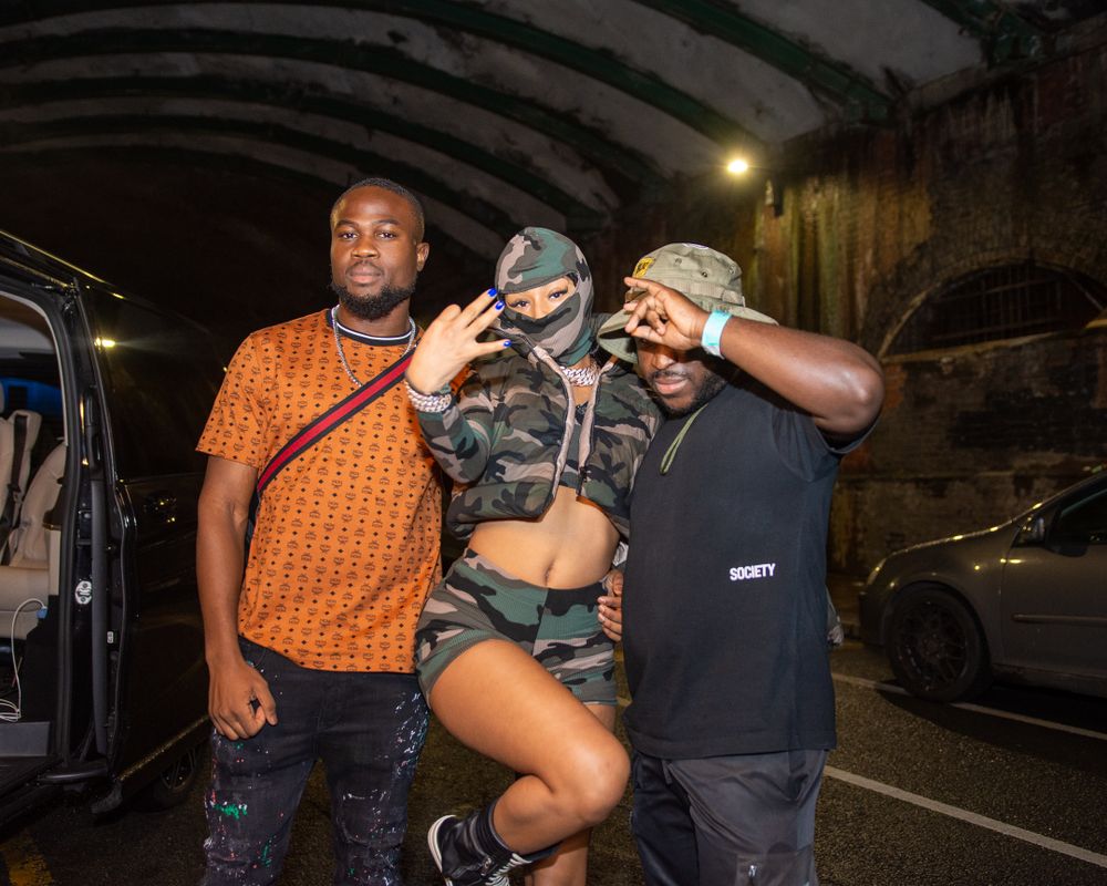 Three friends pose together under a tunnel at night, one wearing a camo mask and crop top.