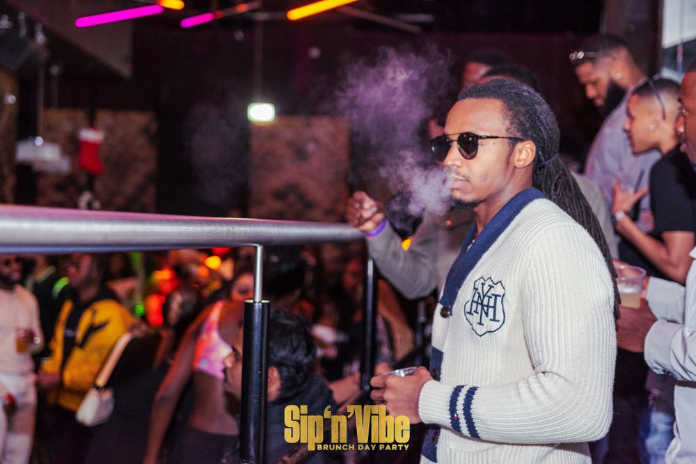 Man with dreadlocks smoking at a lively nightclub party event.