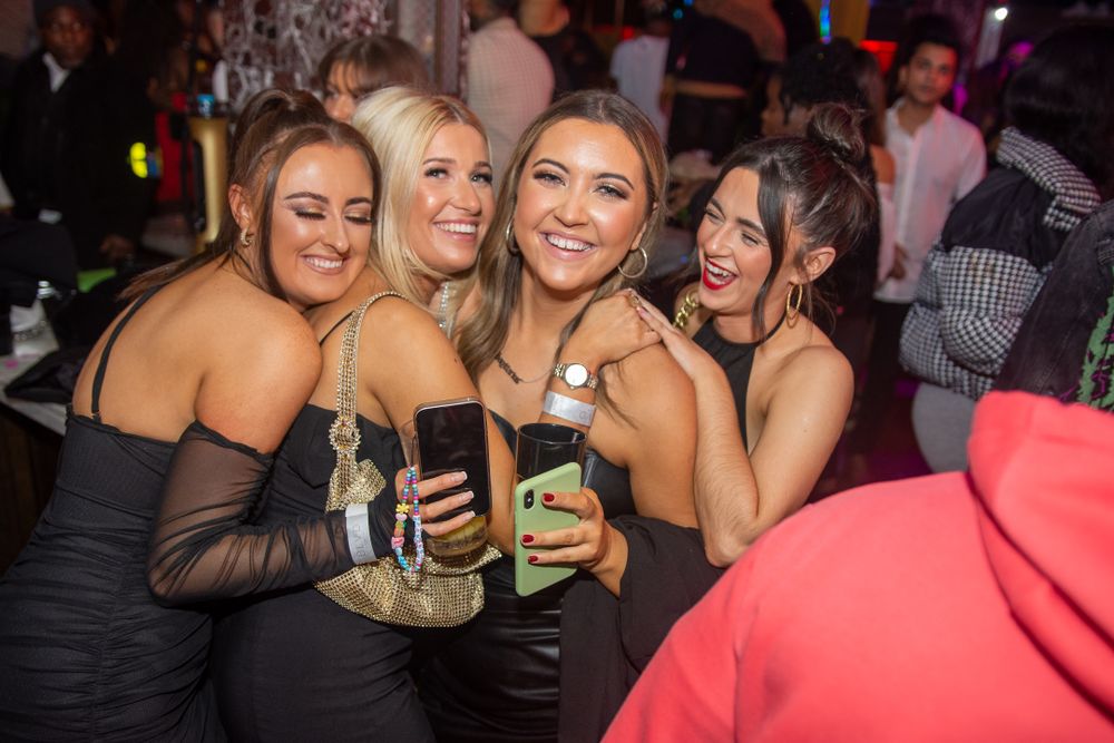 Four women in black dresses smiling and posing together at a nightclub party event.