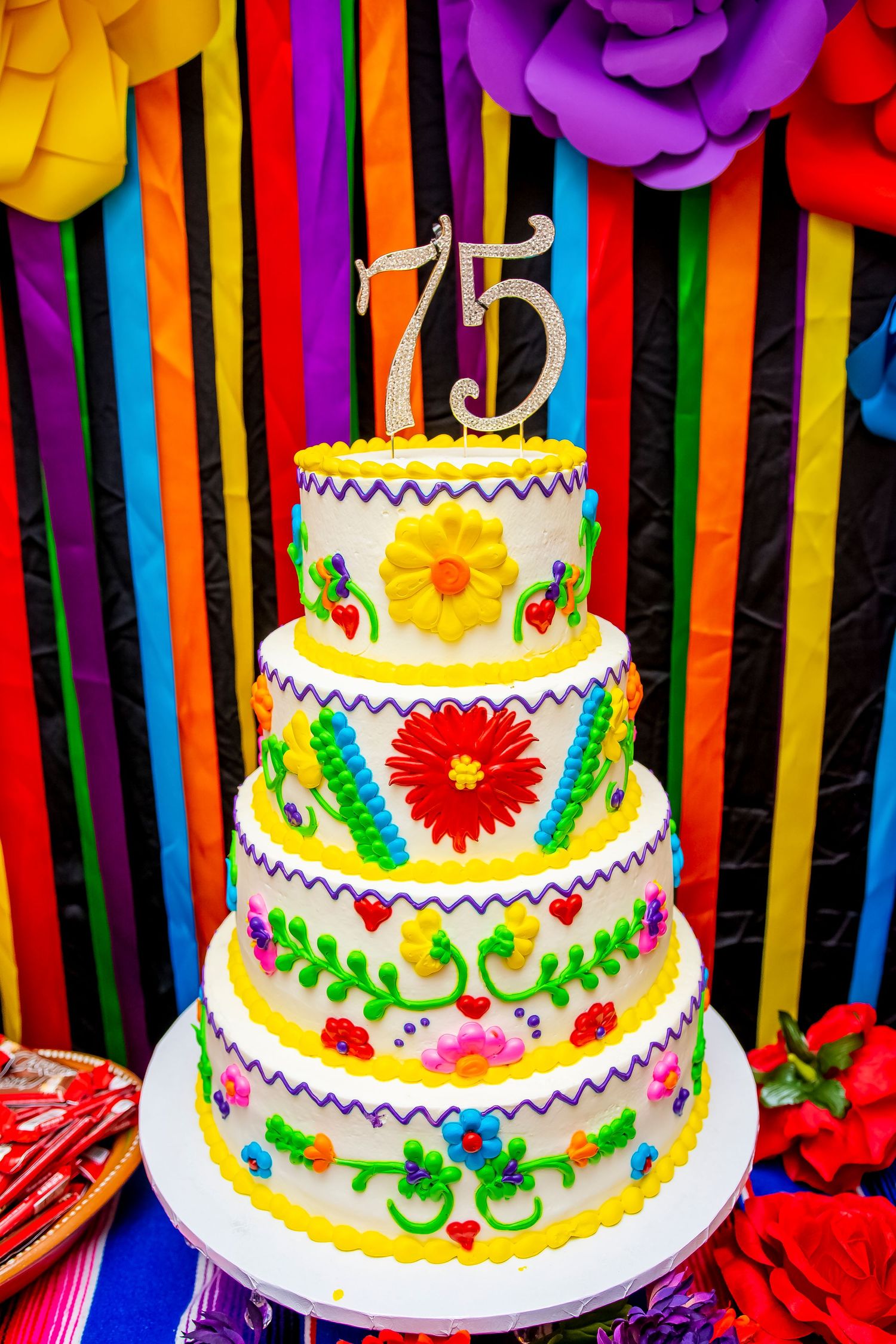 A vibrant tiered birthday cake with '75' topper against colorful striped backdrop with paper flowers.
