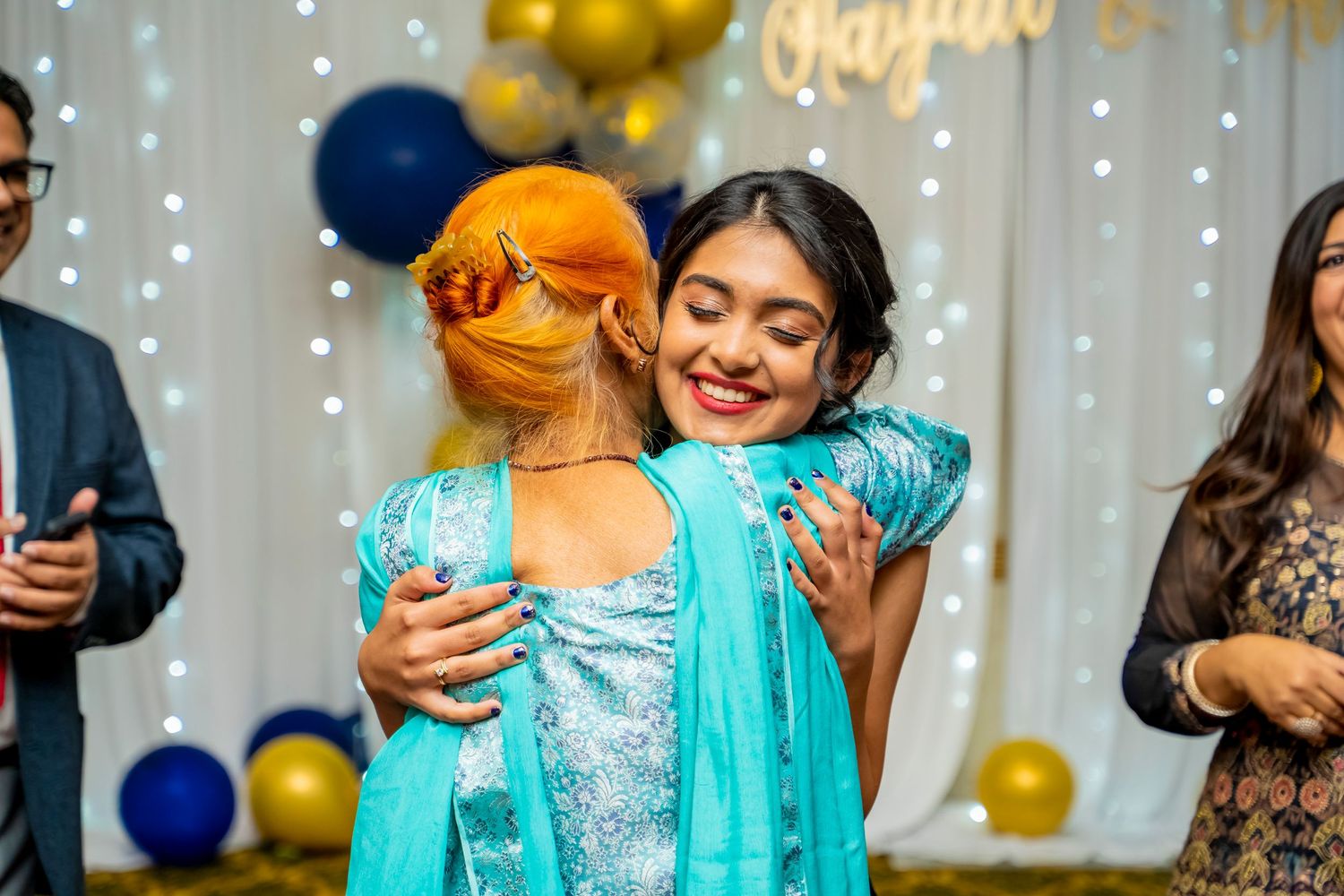 Guest hugging a person in a blue dress at a party with balloons and string lights.