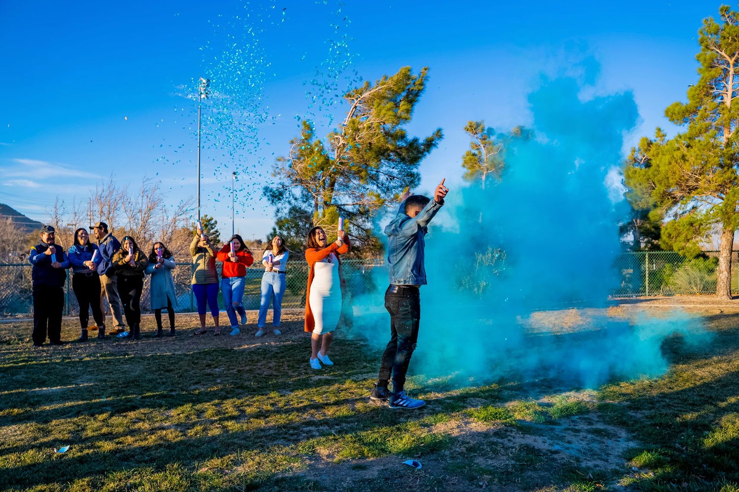 Outdoor gender reveal celebration with blue smoke explosion against clear sky.