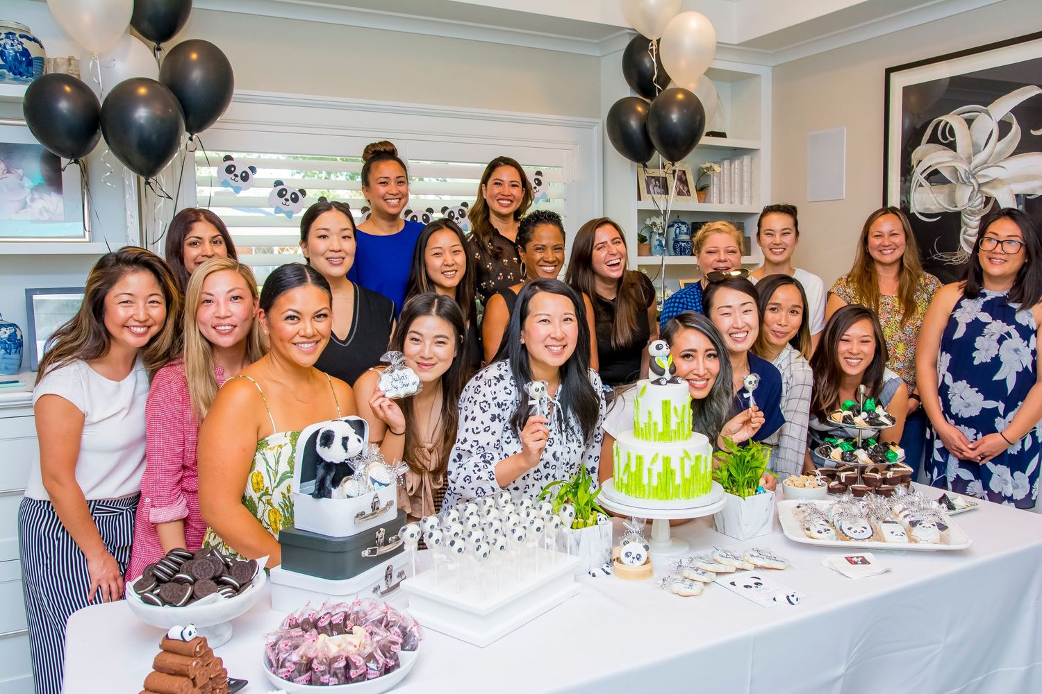 A large group gathers around a festive green and white panda-themed cake with black balloons.
