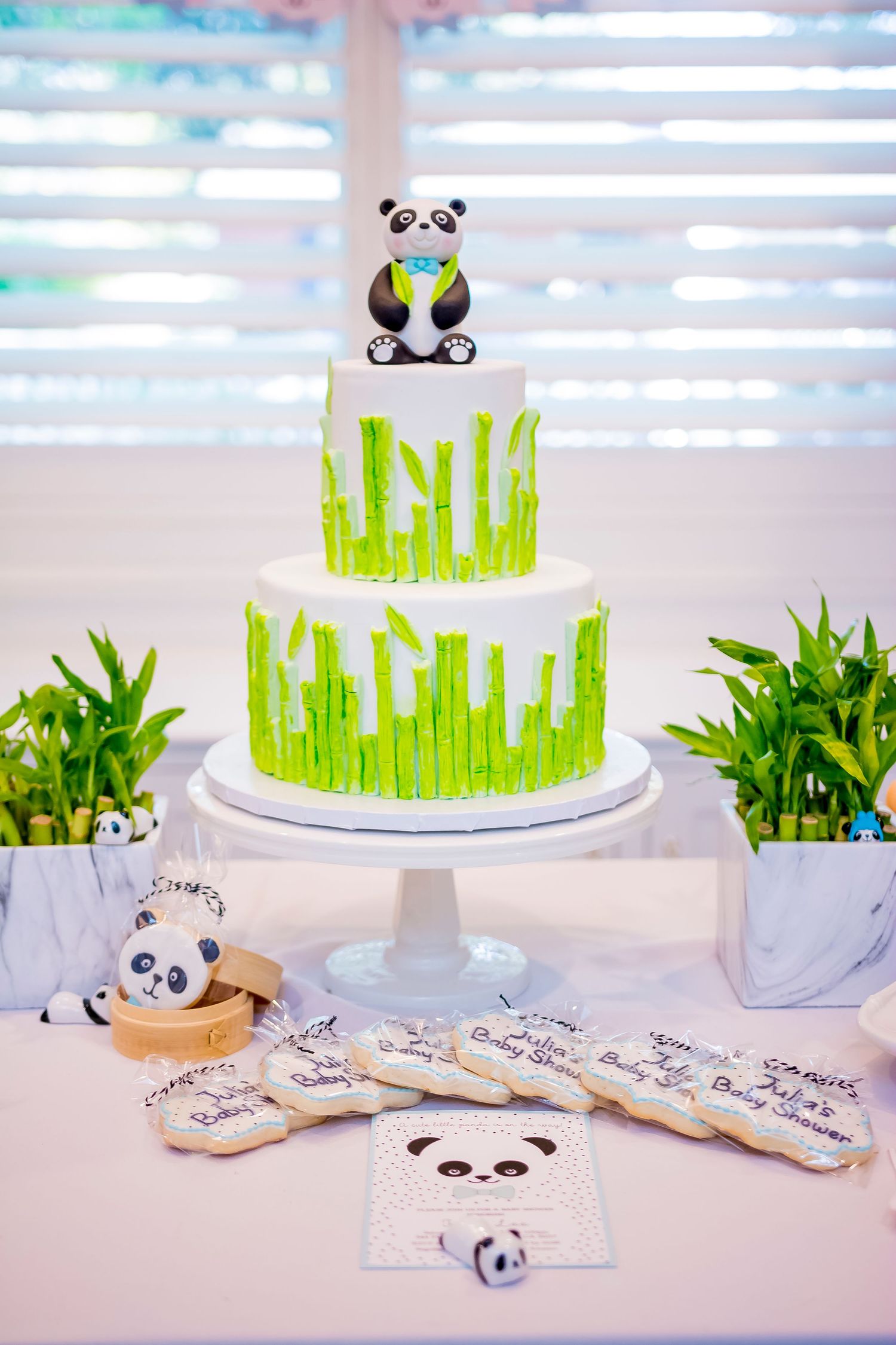White tiered cake decorated with green bamboo designs and a cute panda topper on a marble table.