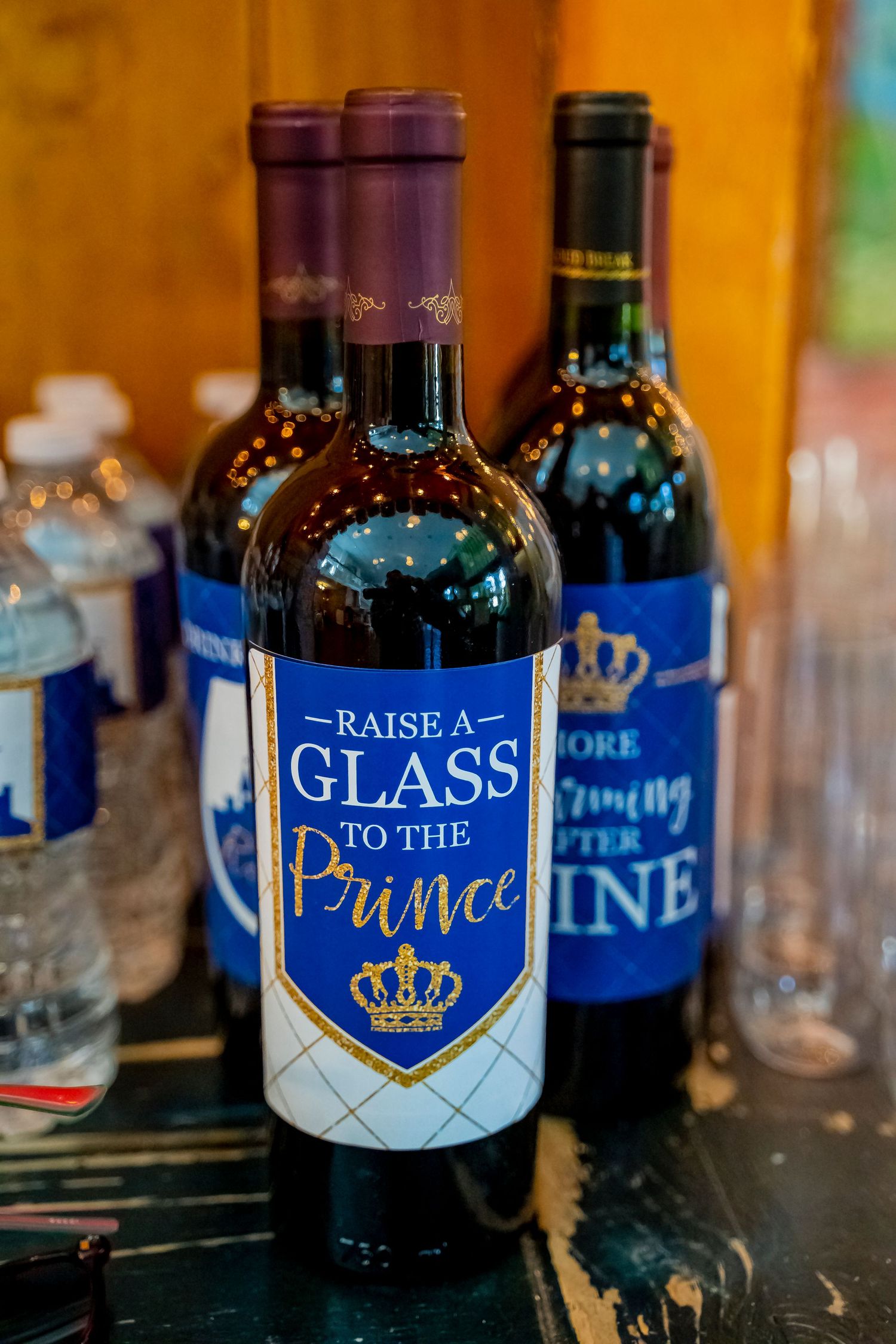 A wine bottle with blue and gold label reading 'Raise a Glass to the Prince' sits among other bottles.