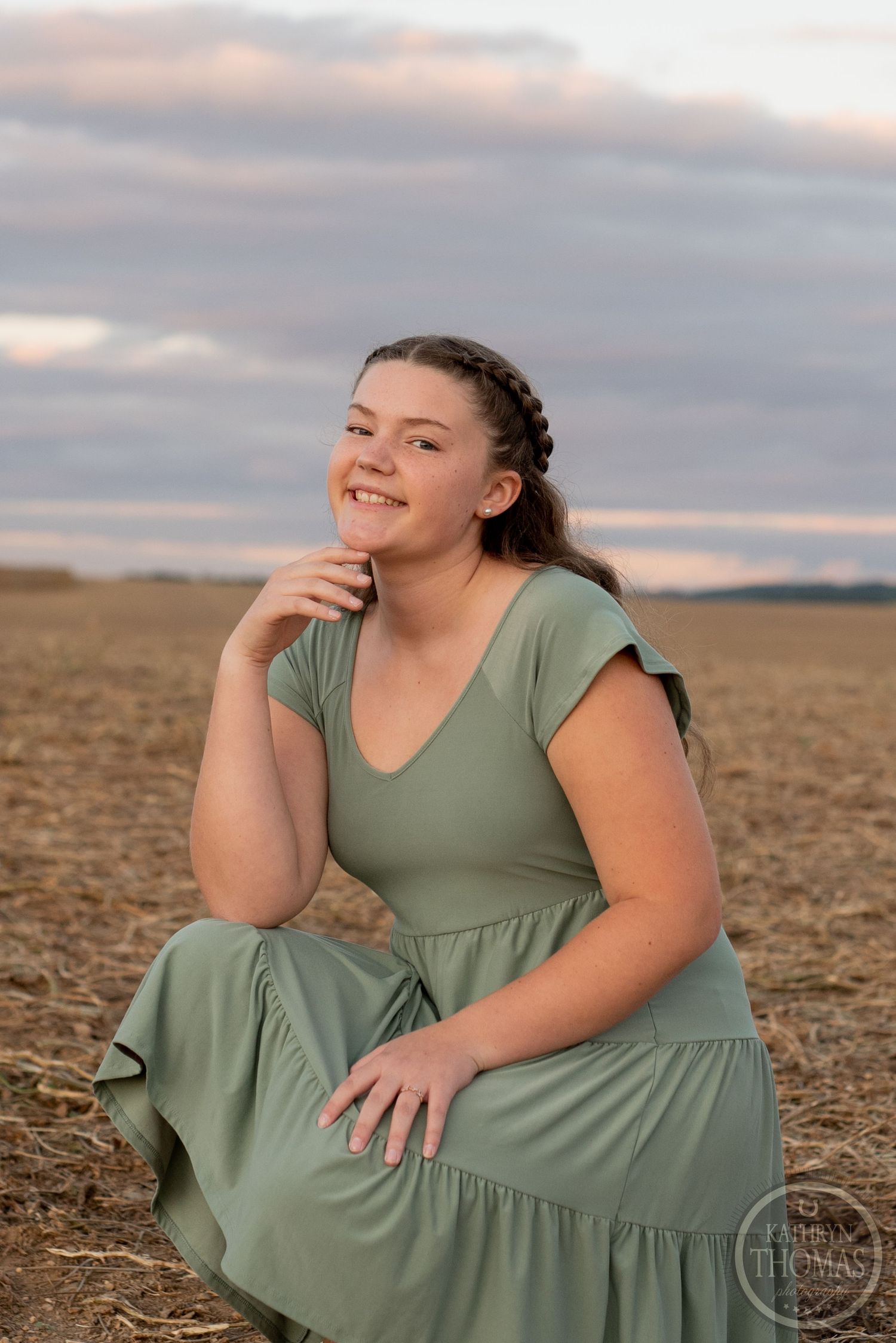 Portrait in a sage green dress against a dramatic sunset sky in an open field.