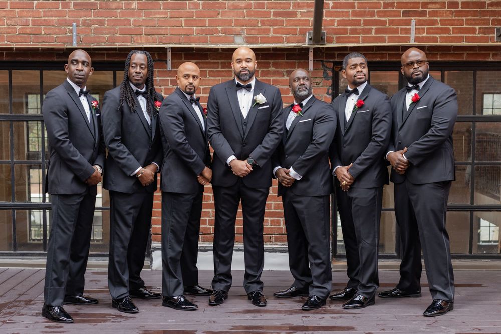 Groomsmen lineup in black tuxedos with red accents against rustic brick wall.