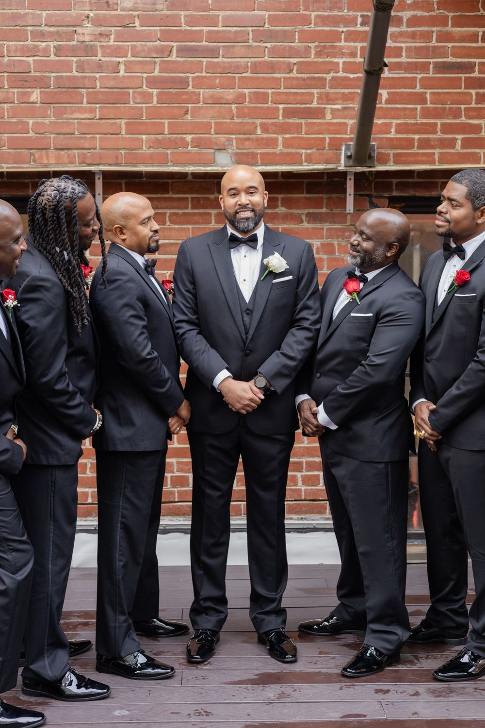 Wedding party members in black tuxedos gather for candid moment against brick backdrop.
