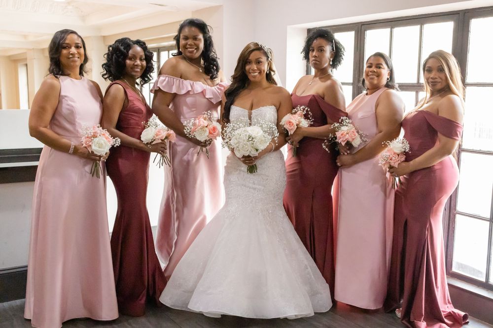 Bridal party poses by window wearing alternating pink and burgundy floor-length gowns.