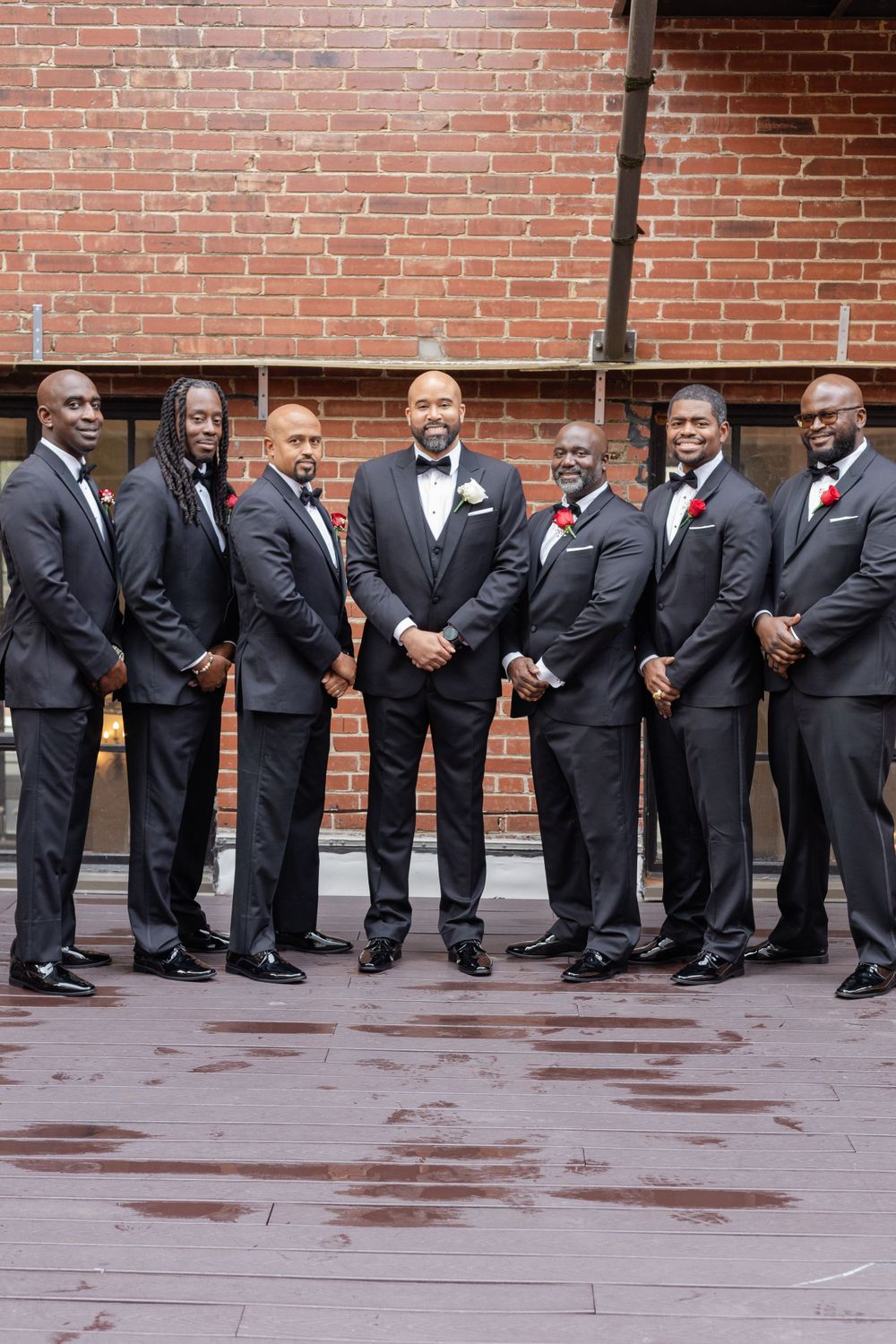 Groomsmen in black tuxedos with red boutonnieres pose against brick wall.