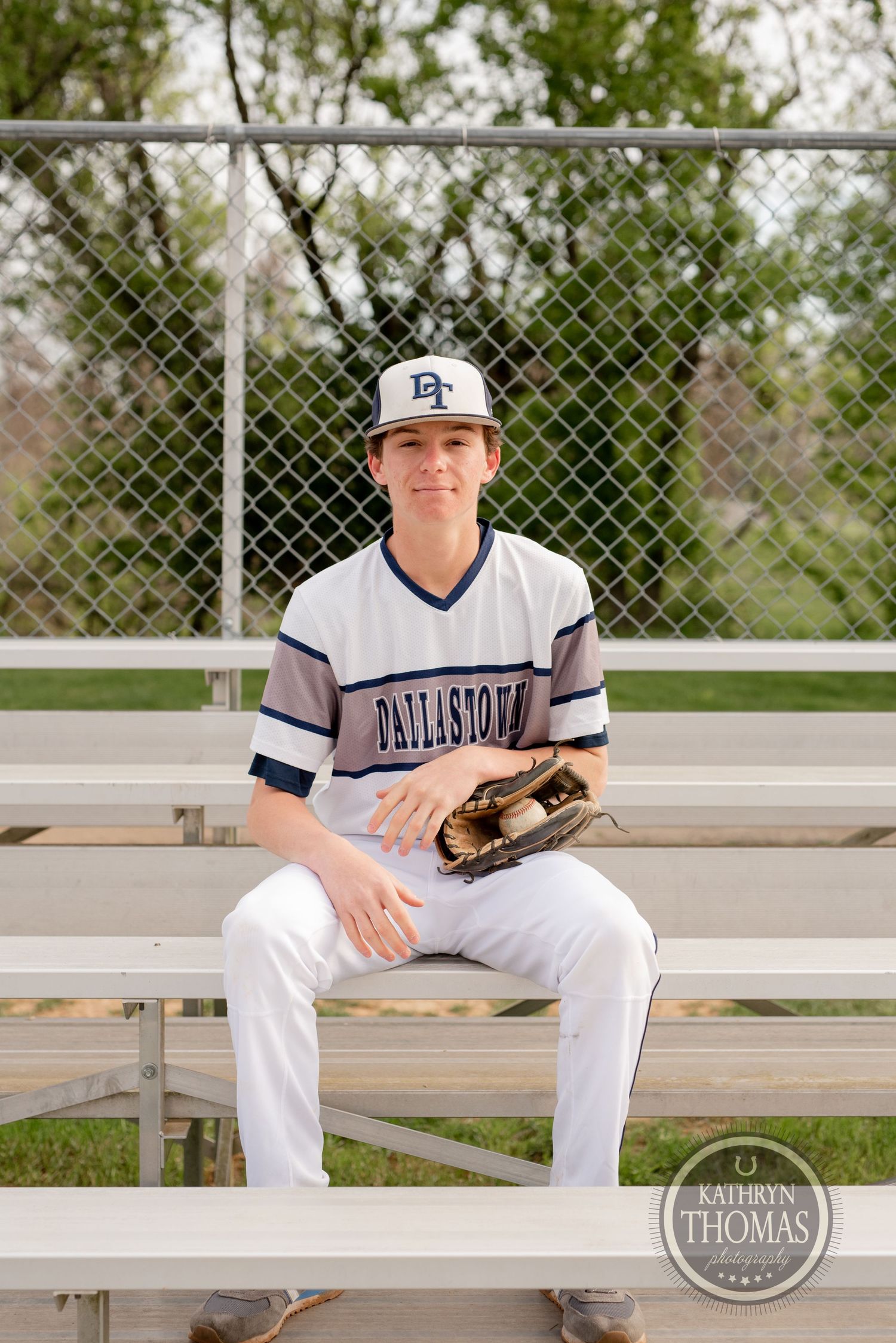Baseball player sits on bleachers with glove wearing white Dellasoft uniform.