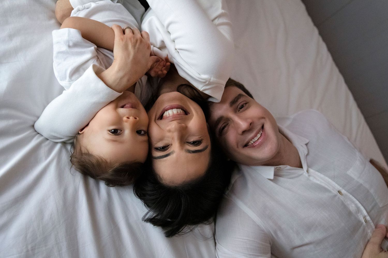 A happy family lying together on white bedding sharing genuine laughter and smiles.