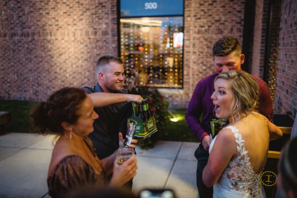 Wedding guests share drinks and laughs on an outdoor patio with brick walls and evening lighting.