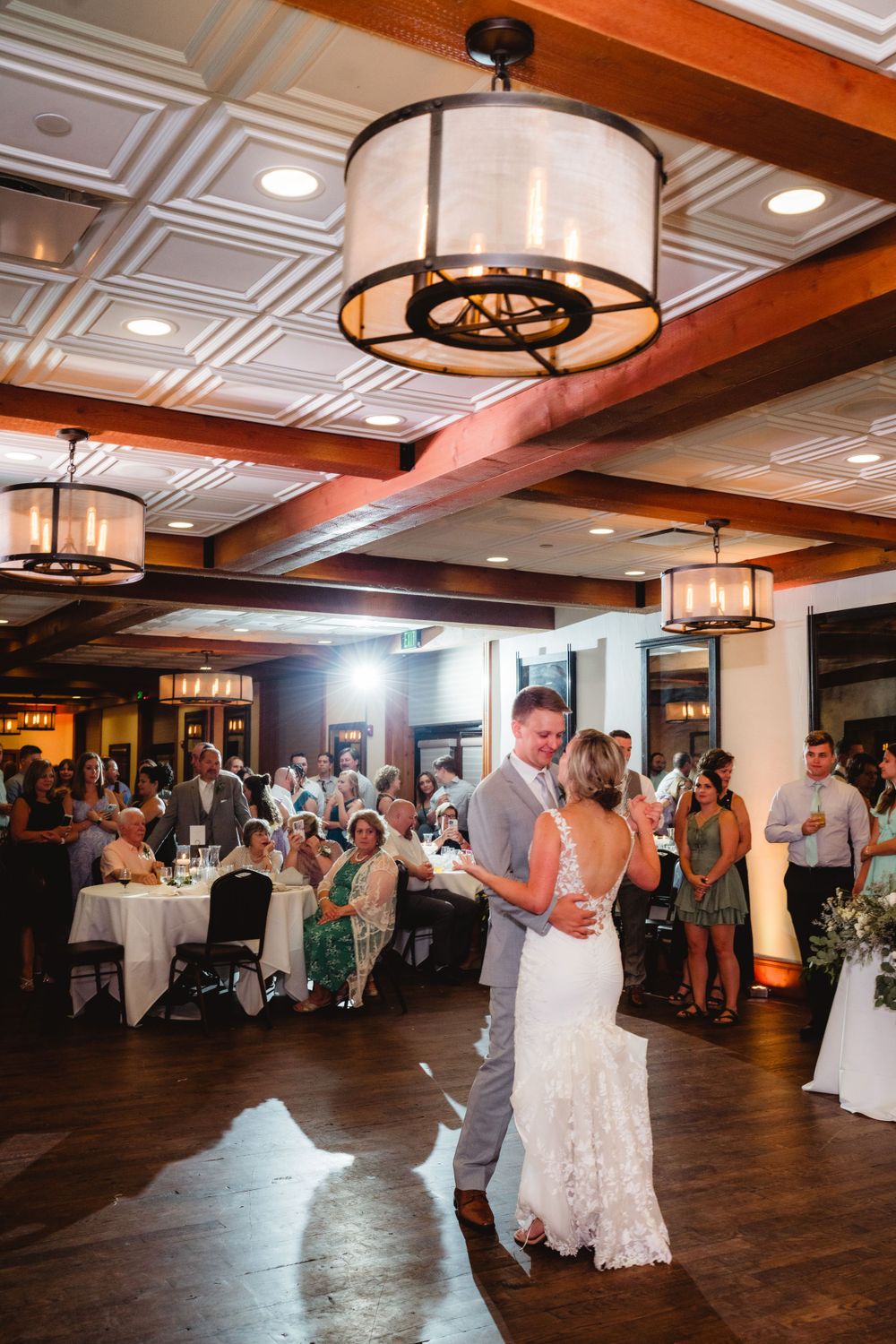 Couple dancing at a wedding reception in an elegant ballroom with wood floors and decorative lighting fixtures.