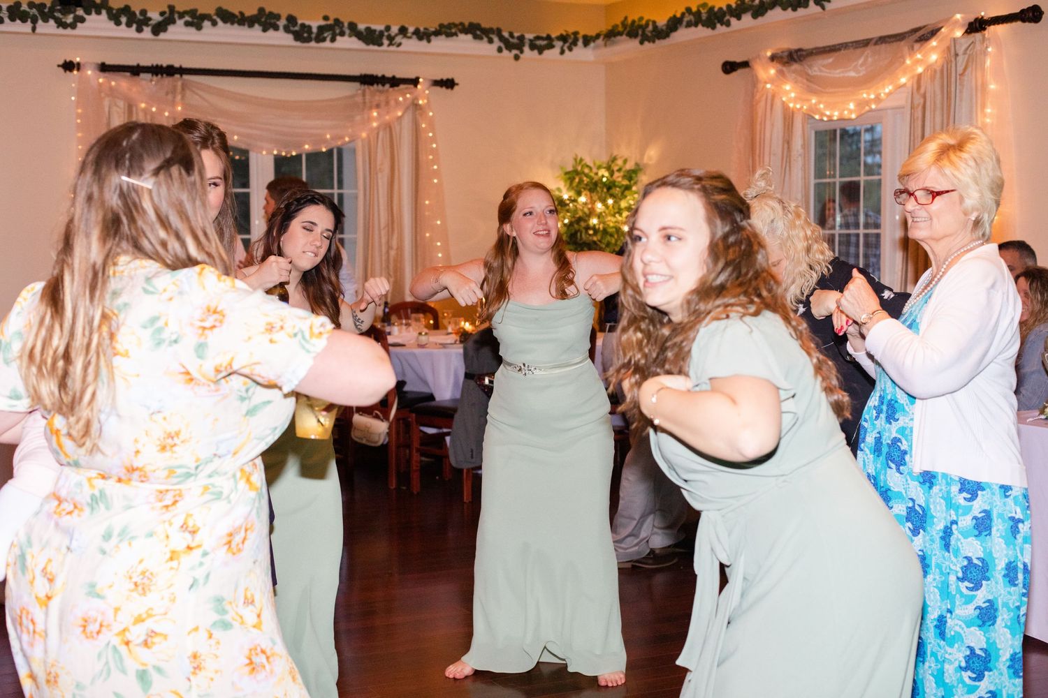 Wedding guests in pastel dresses dancing together at a reception venue.