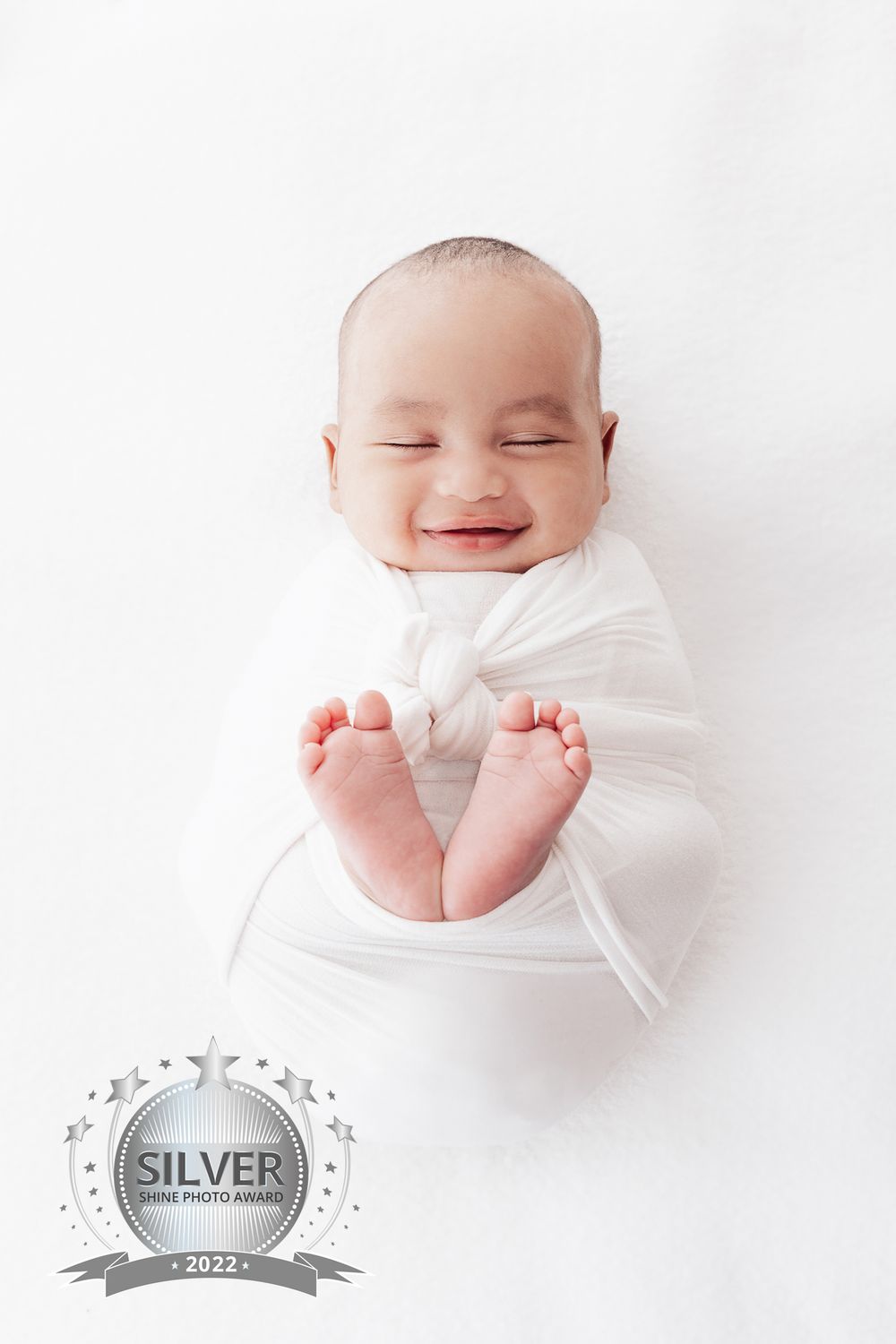 Smiling newborn baby wrapped in white with tiny feet showing against white background.